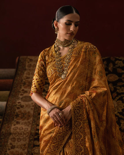 Woman in a traditional orange saree with intricate patterns, standing against a dark background.