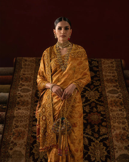 Woman in a traditional gold saree standing against a textured wall.