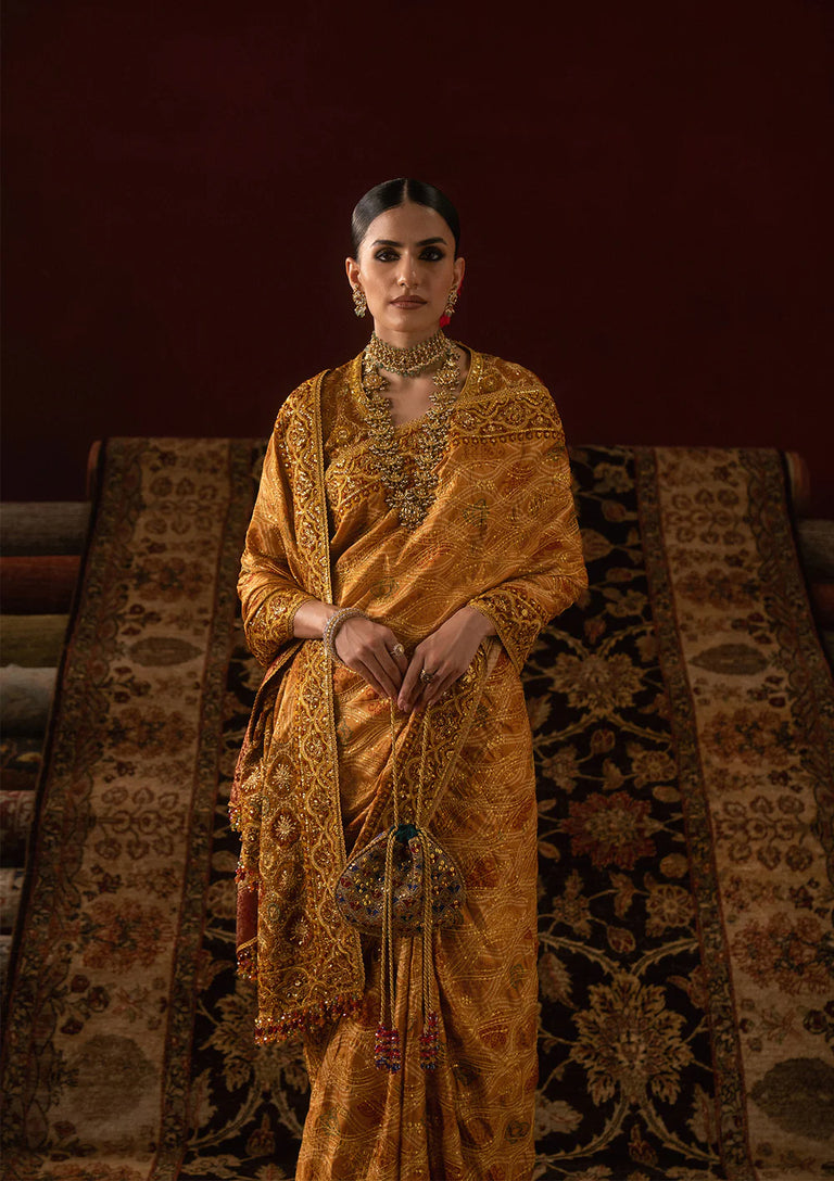 Woman in a traditional gold saree standing against a textured wall.