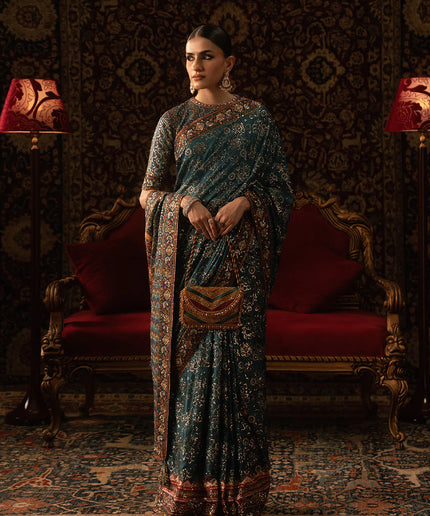 Woman in a traditional saree standing in an ornate room with red chairs and patterned walls.