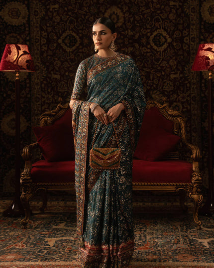 Woman in a traditional saree standing in an ornate room with red chairs and patterned walls.