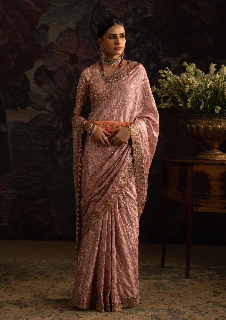 Woman in a traditional pink saree with intricate patterns, standing against a dark background.