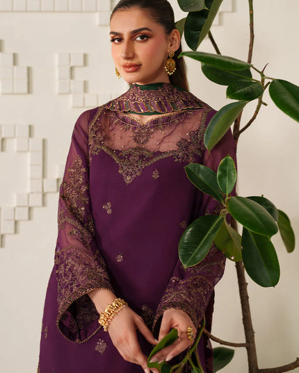Woman in a purple traditional outfit standing next to a plant against a white tiled wall.